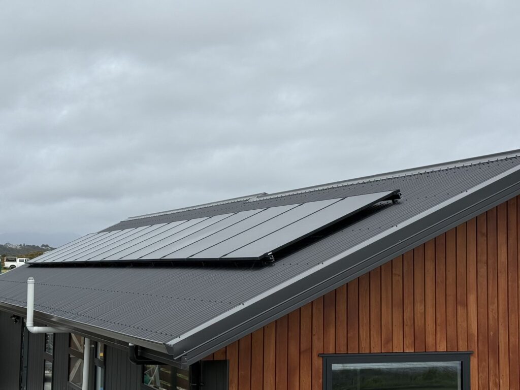 A set of black solar panels is installed on the slanted metal roof of a wooden building under a cloudy sky.