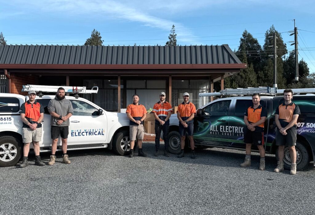 Seven men in work uniforms stand in front of two service vehicles with company logos, parked outside a building with a corrugated metal roof.