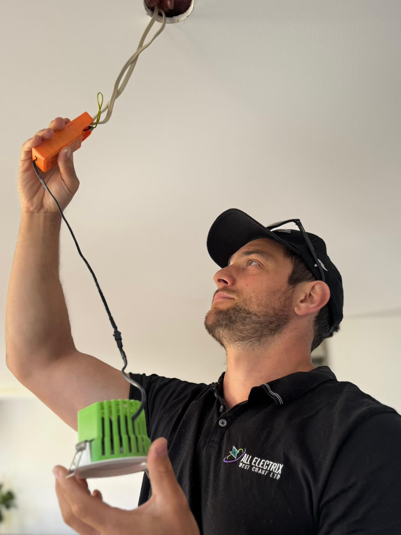 A man in a black shirt and cap tests electrical wiring on a ceiling while holding a green and white device and an orange tool.